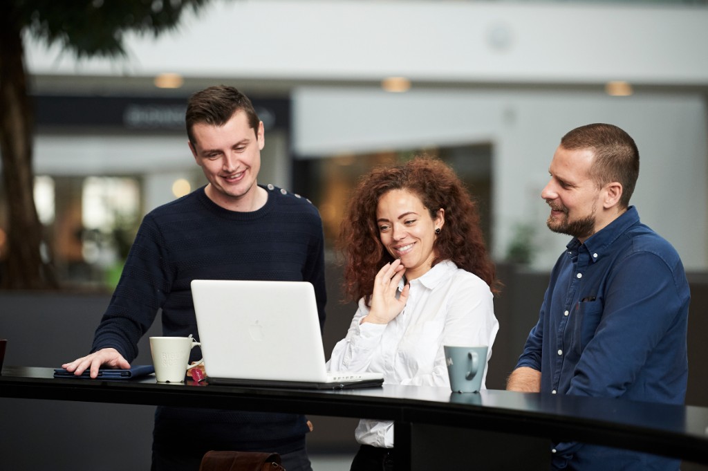 Three teammates around a laptop at a high-top table — bright atrium-style office, shallow depth of field.