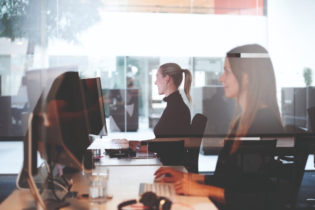 Desks and people in a glass-partitioned office — high-key light, documentary workplace feel.