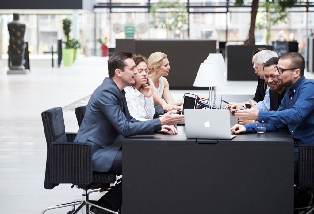 Six people in discussion at a long black table — large windows, soft natural light.