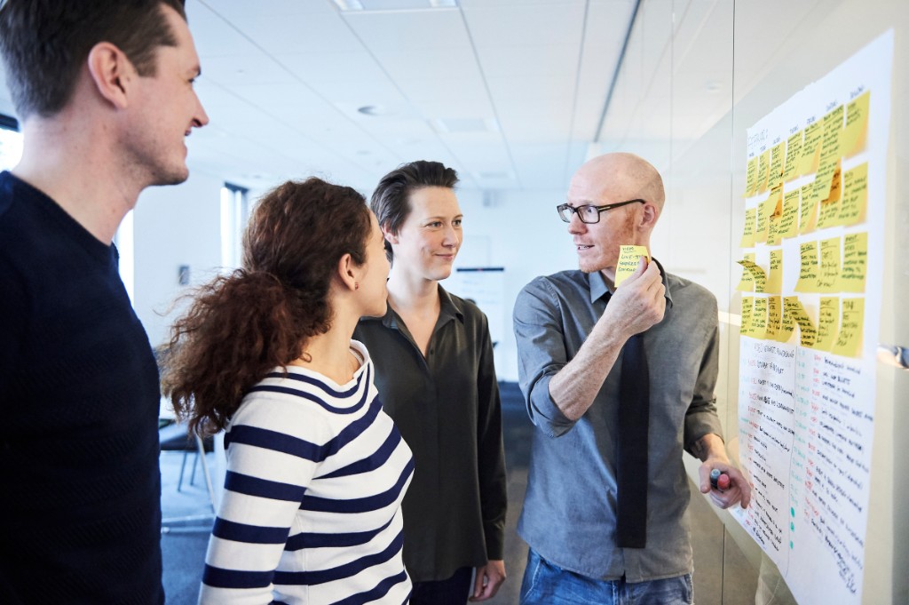 Team at a glass wall with yellow sticky notes — agile planning, cool neutrals with a colour accent.