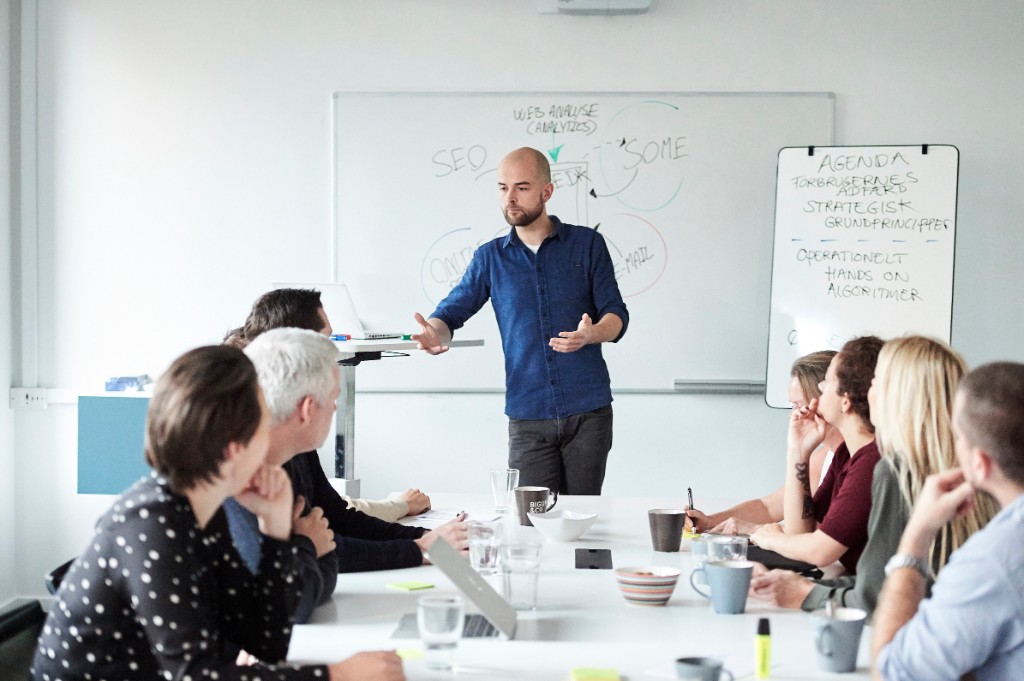 Workshop: presenter at the head of a long table with whiteboards — bright, candid training room.
