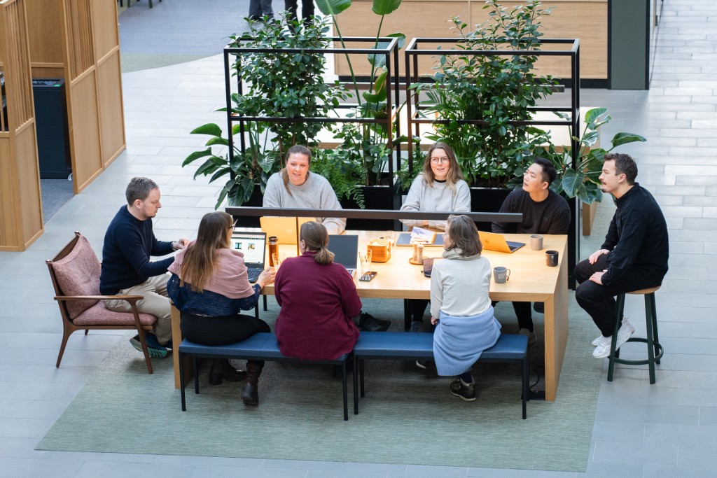 Bird’s-eye view of a team around a communal oak table — plants, muted seating, Scandinavian modern.