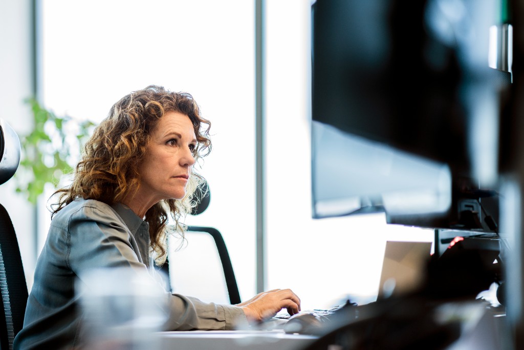 Woman working at a desk in profile — blurred monitor foreground, bright window light, shallow depth of field.