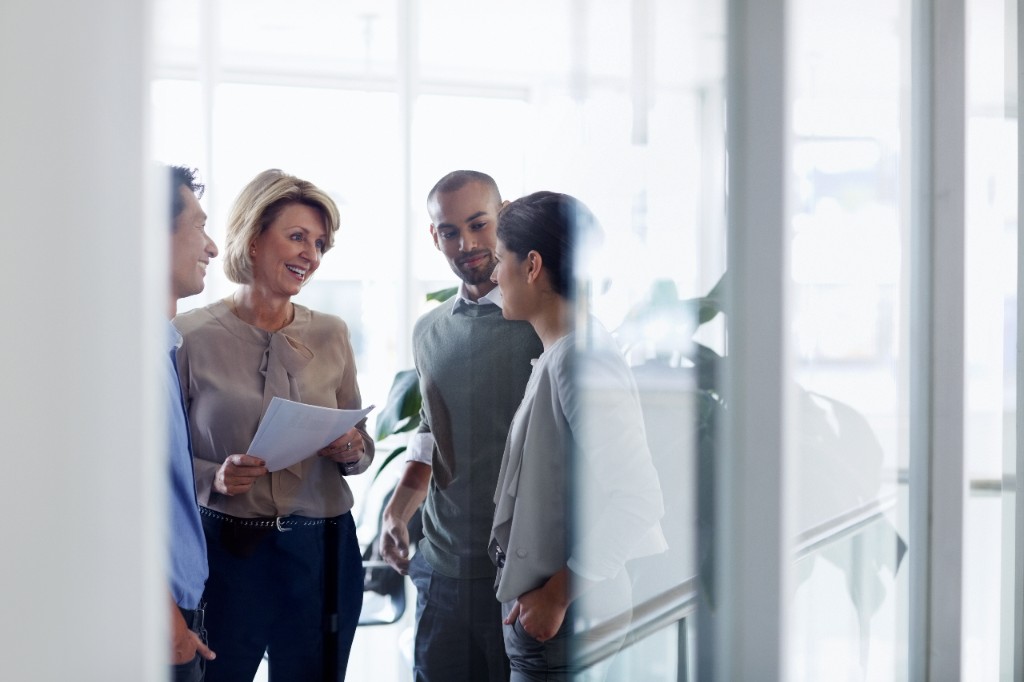 Four colleagues in conversation photographed through glass — reflections, airy modern office.