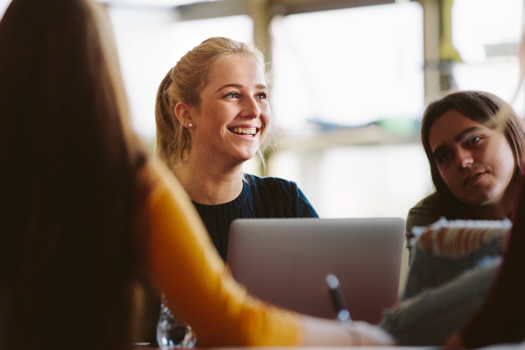 Young woman laughing at a collaborative table — peers softly blurred around her, laptop in frame.
