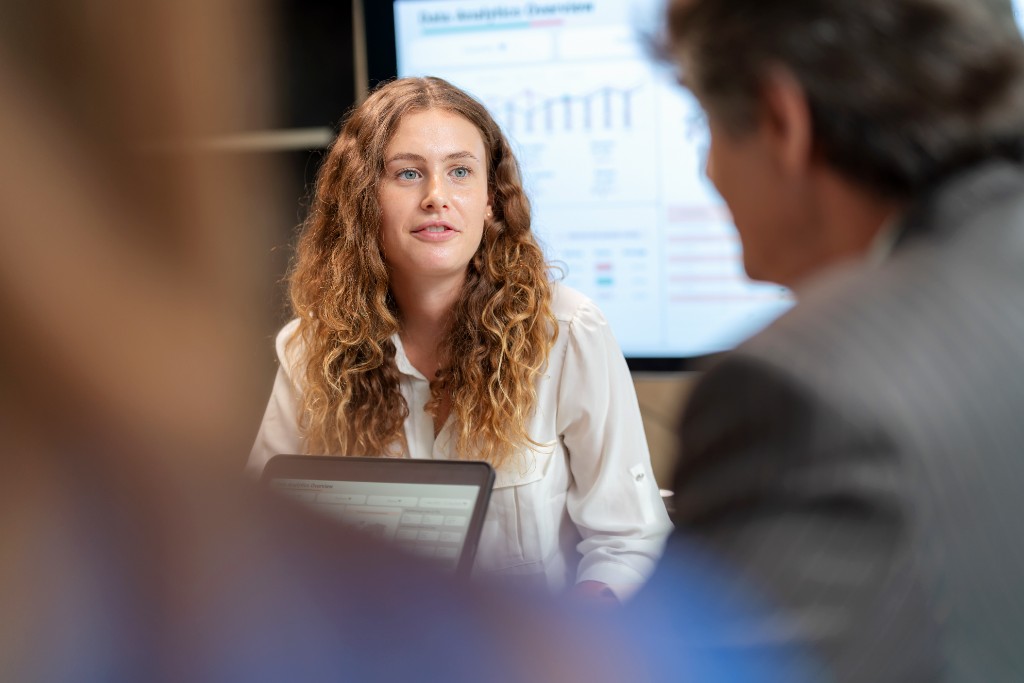 Collaborative dialogue with analytics on a conference screen — layered foreground shoulders, cinematic bokeh.