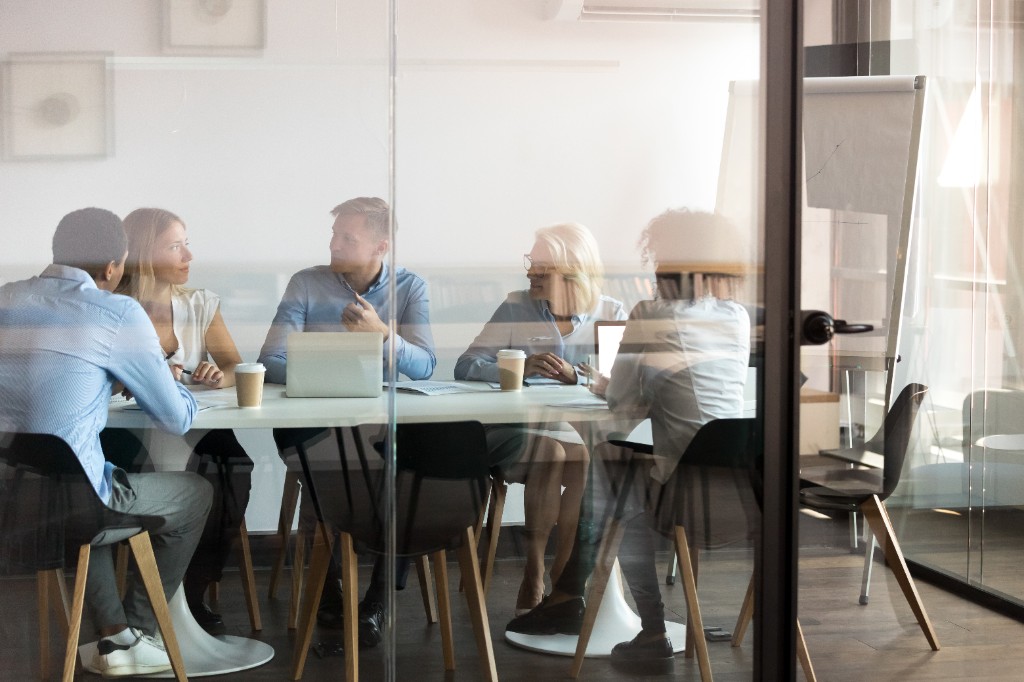 Meeting in a glass-walled conference room seen from outside — candid teamwork, minimalist furniture.