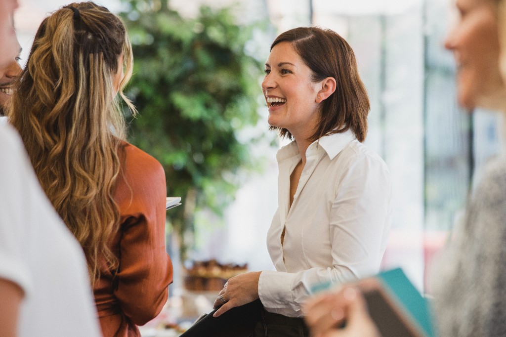 Small group conversation — centred laugh, leafy plant behind, soft natural light.