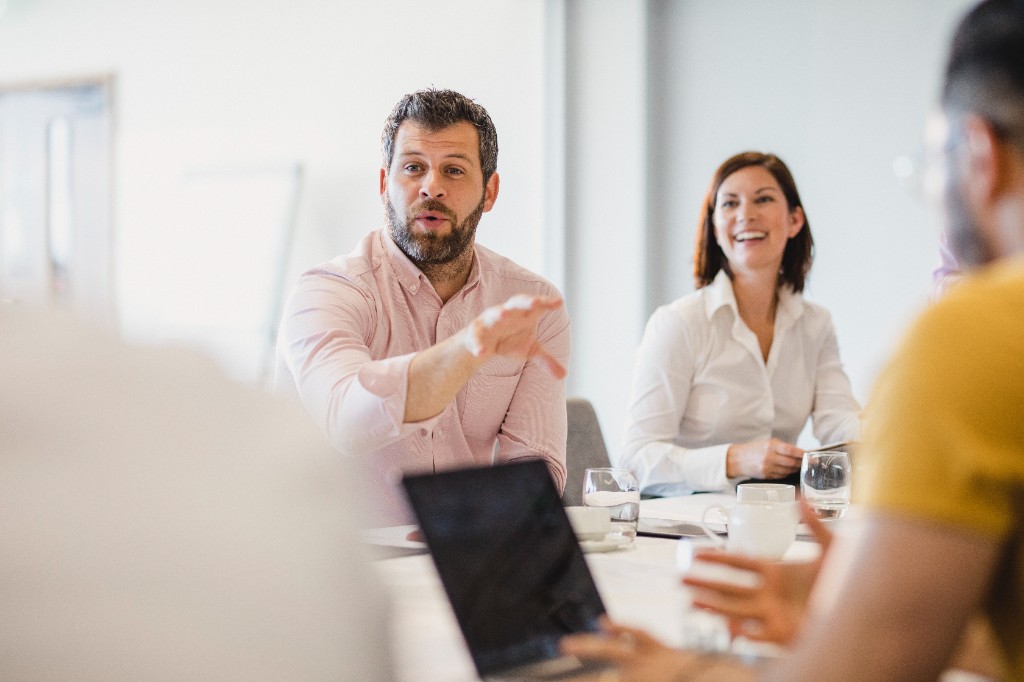 Colleague gesturing mid-conversation beside a smiling teammate — laptops and cups on a white table.