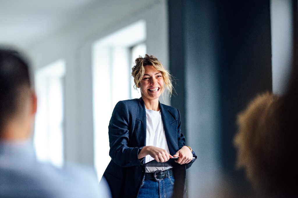 Business-casual professional laughing amid peers — denim and navy blazer, blurred foreground silhouettes.